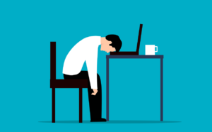 Employee sitting on a chair in front of a table with a laptop and a coffee mug on it. The employee's head is faced down with his forehead on the keyboard. This picture is intended to act as an example as a stressful moment, hence why the caption is made up of the word "Stress"