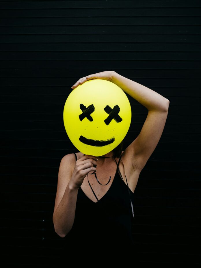 Person standing in front of black backdrop, holding a yellow happy face balloon with x's for eyes, to cover their face