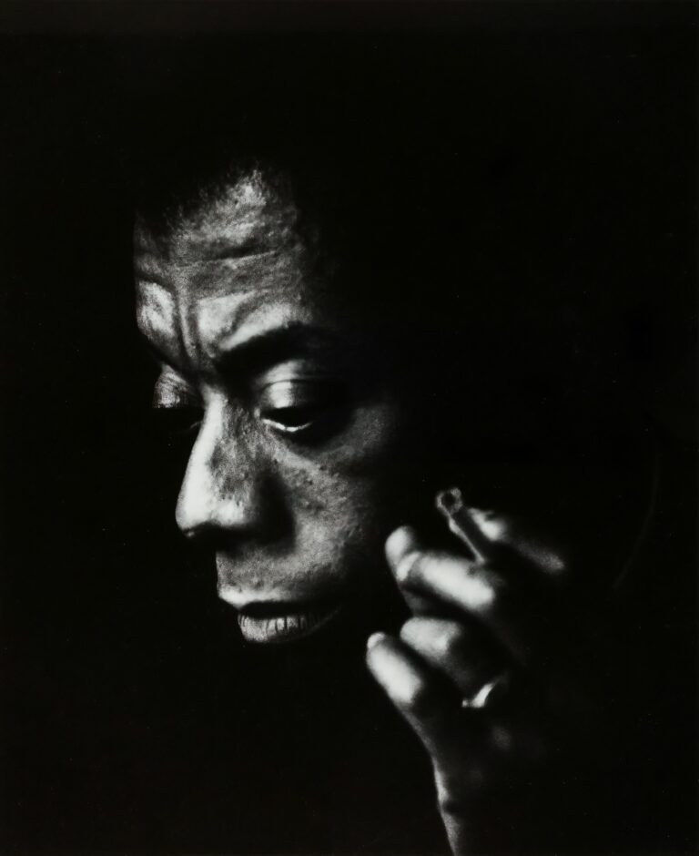 Black and white headshot of author James Baldwin holding a cigarette. Netherlands, 1965