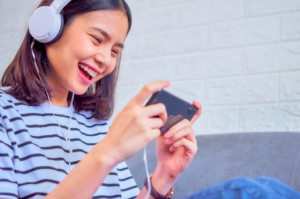 Excited young Asian woman sitting on sofa wearing white headphone on the head and playing games on smartphone...